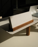 A Magnet Kit, featuring a pen tray and an Analog Weekly calendar with blank weekly pages, rests in its wooden holder atop a Gather Monitor Stand on a modern desk bathed in sunlight.