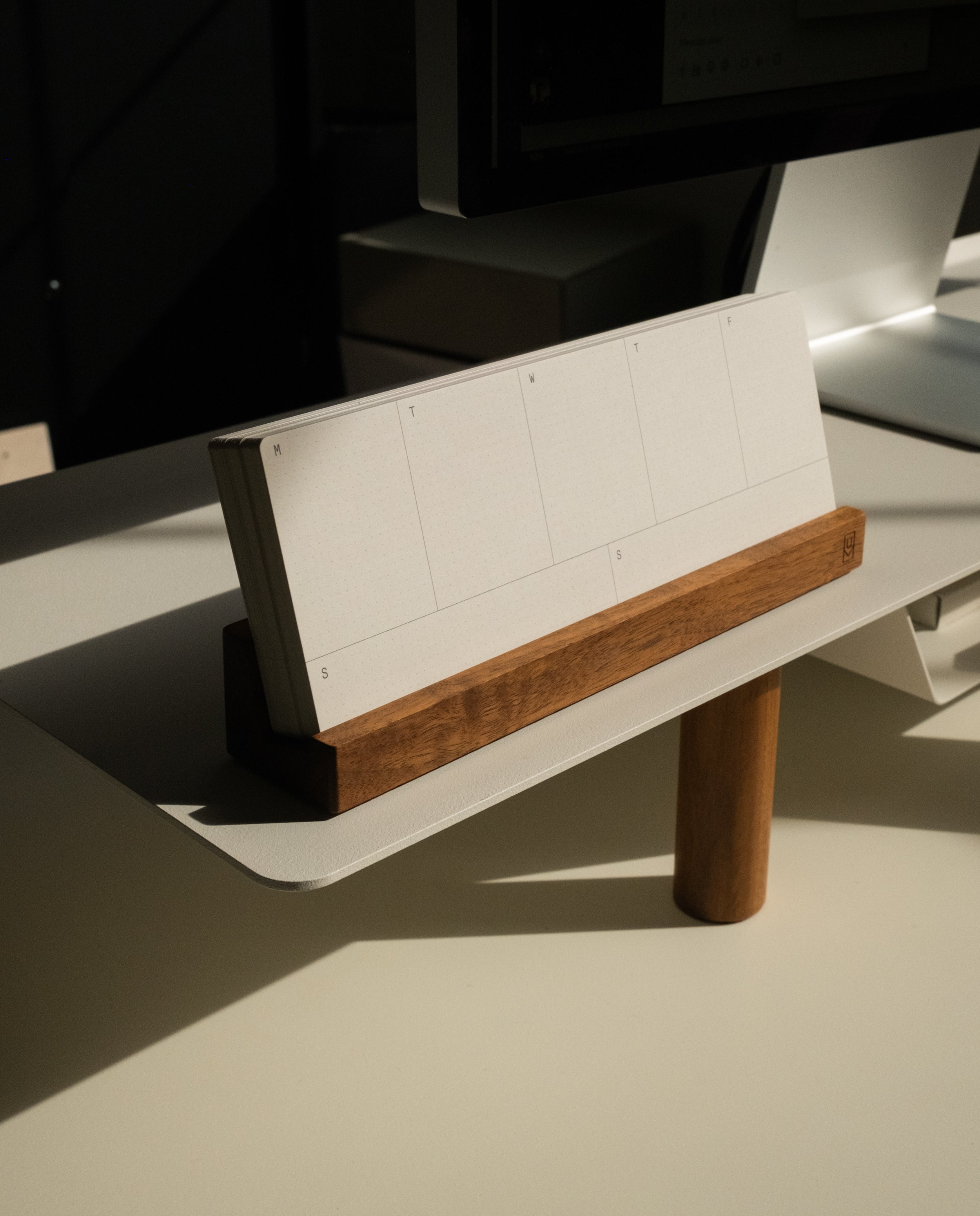 A Magnet Kit, featuring a pen tray and an Analog Weekly calendar with blank weekly pages, rests in its wooden holder atop a Gather Monitor Stand on a modern desk bathed in sunlight.