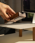 A hand places the Magnet Kit (Pen Tray and Analog Weekly) onto a light-colored desk with wooden legs, with a computer monitor visible in the background.