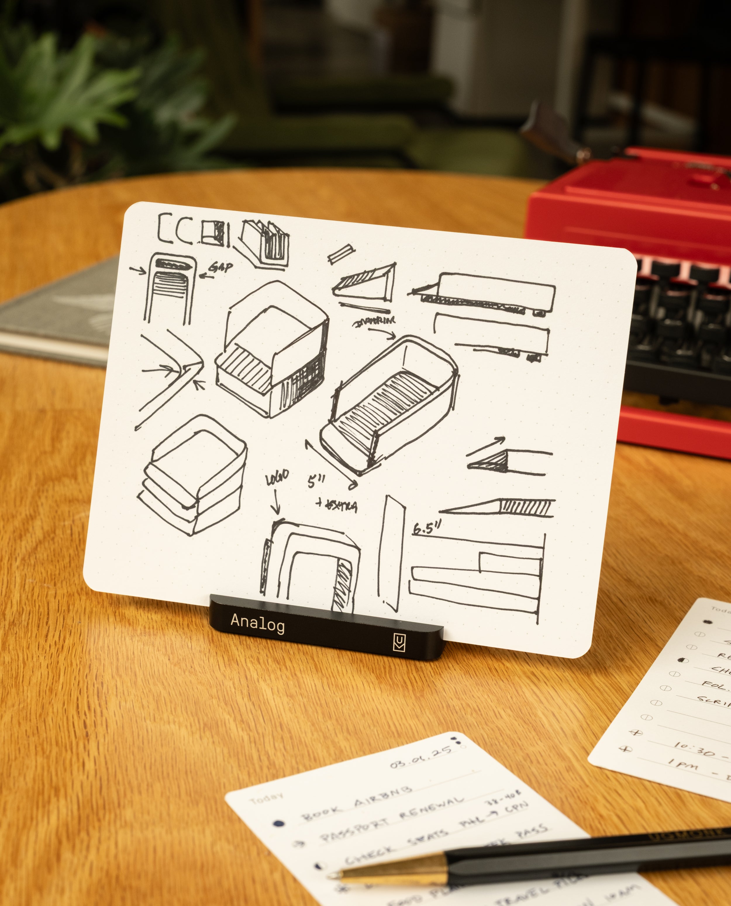 An Analog Starter Kit (Black) card—featuring black ink sketches of stackable containers—rests in its aluminum card stand on a desk beside a red typewriter, a plant, and handwritten notes, evoking the charm of a paper card system.