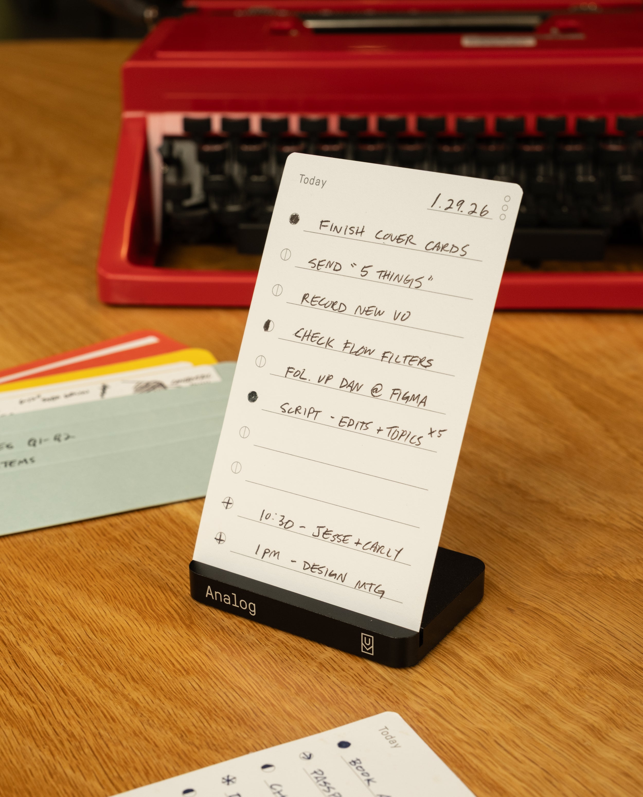 A handwritten daily task list from the Analog Starter Kit (Black) is displayed on an aluminum card stand atop a desk, with a red typewriter and file cards visible in the background. The list, organized through this paper card system, features tasks such as recording VO and editing scripts.