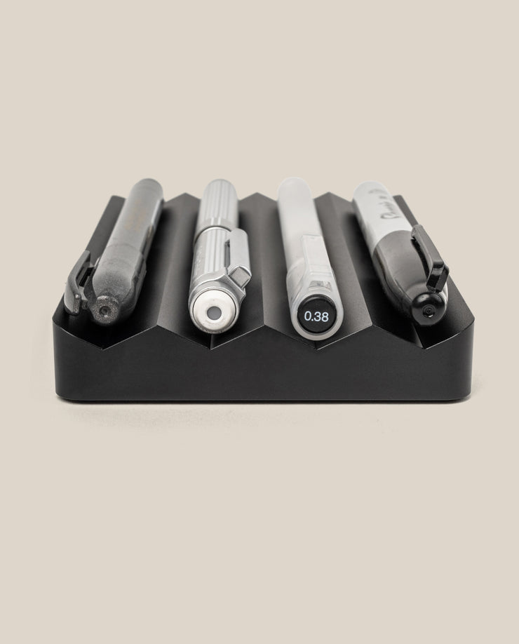The Complete Analog Collection (Black Aluminum), featuring four uniquely designed pens, is displayed horizontally in individual grooves of a black desktop organizer against a plain beige background.