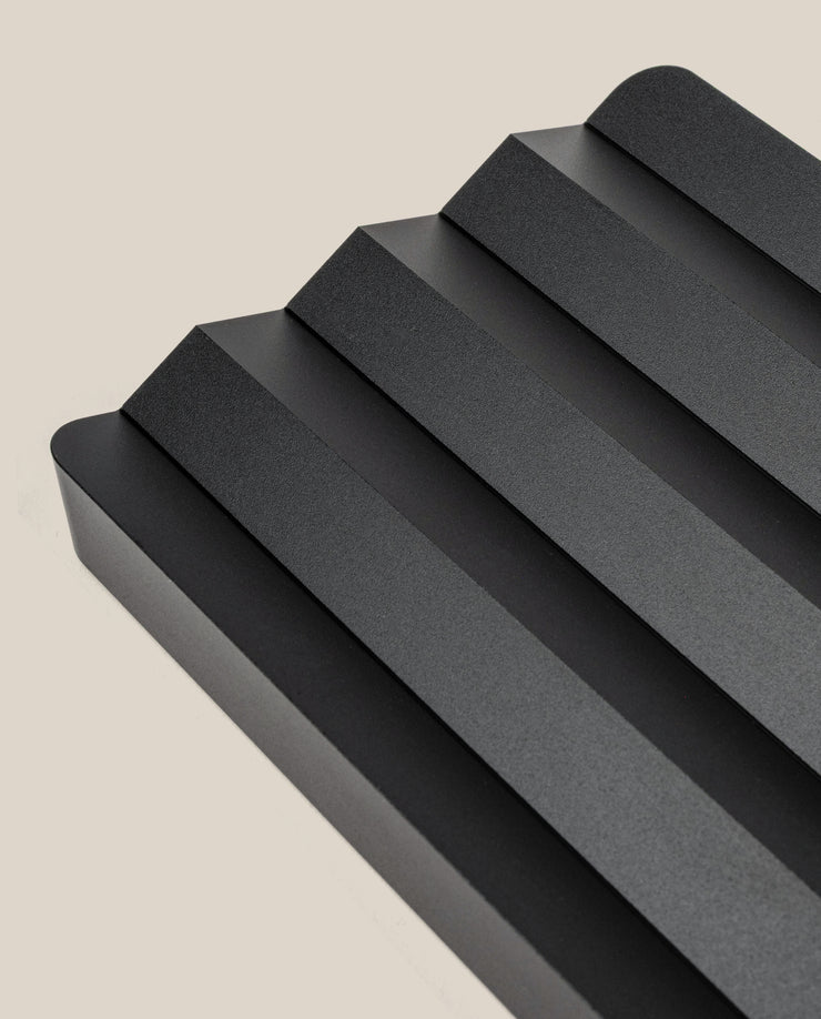 The Complete Analog Collection (Black Aluminum) features a sleek, minimalist tray with four angled grooves and rounded edges, photographed on a light beige background—a modern geometric desktop organizer.