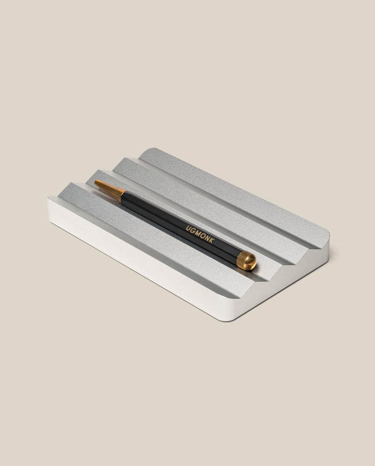 A black and gold pen with UGMONK branding rests on the Analog Multi Pen Tray (Silver Aluminum) against a light beige background.
