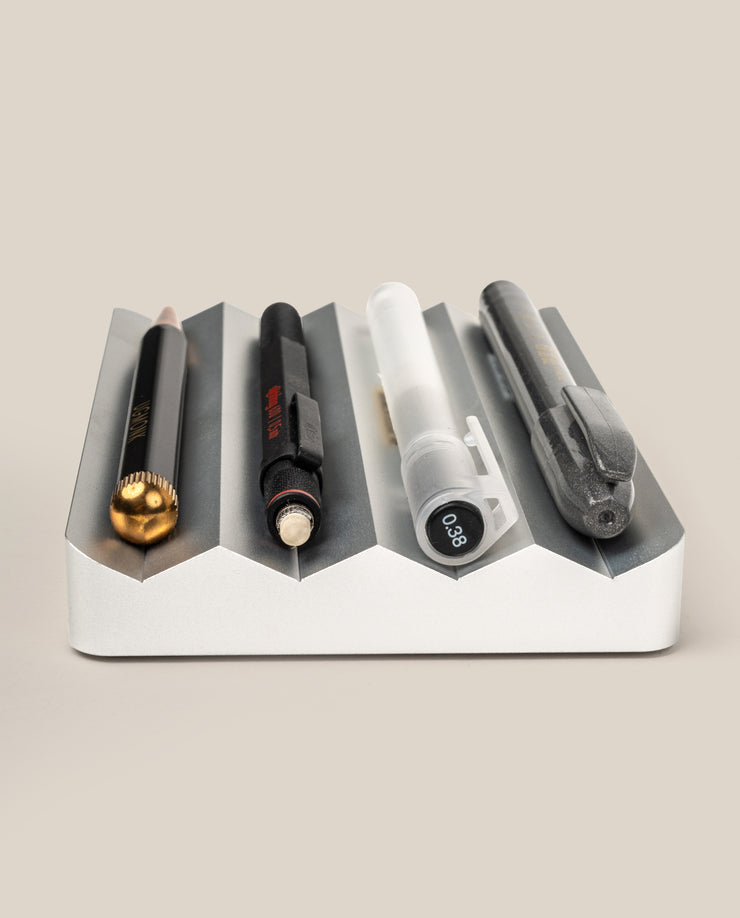 The Complete Analog Collection (Silver Aluminum) desktop organizer, shown on a light beige background, holds four writing tools: a black and gold pencil, a black pen, a translucent mechanical pencil, and a gray pen.