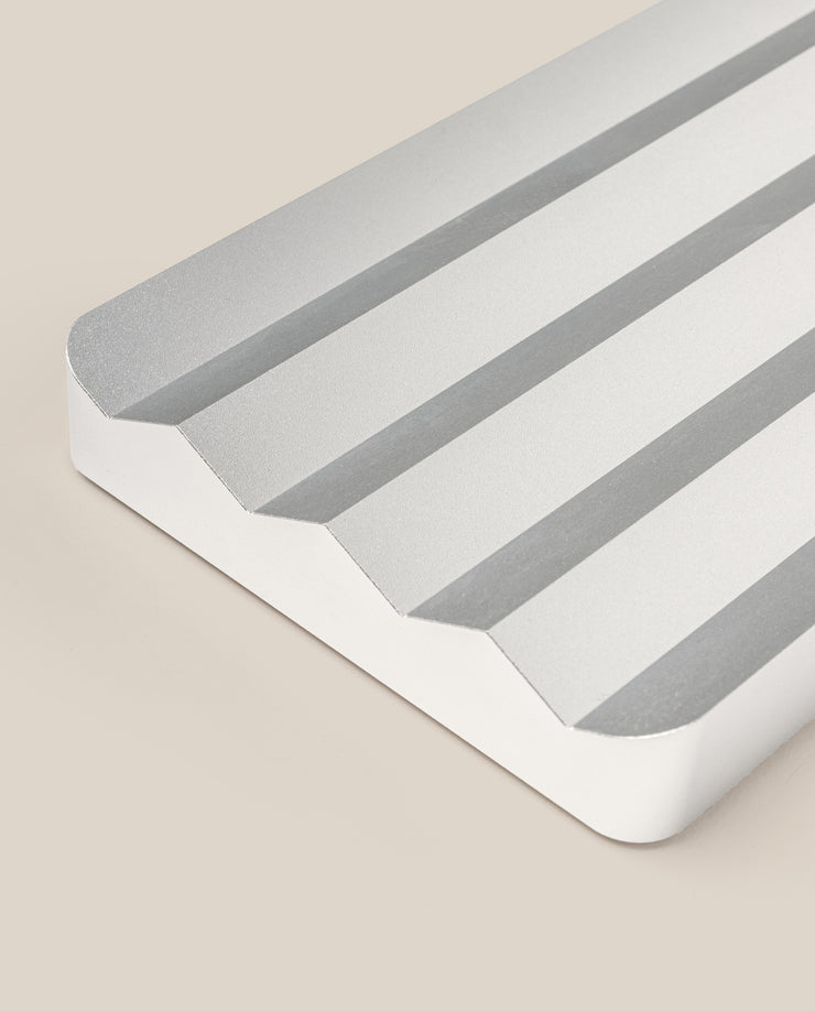 A close-up of the Analog Multi Pen Tray (Silver Aluminum), a sleek rectangular pen holder with three angled grooves, white sides, and rounded corners, displayed on a beige background.