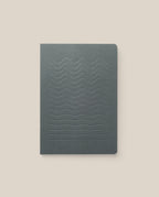 The Layflat Notebook (Charcoal) features a dark gray cover with embossed wavy lines, a minimalist design, dot grid pages, and Smyth sewn binding. It is shown on a light beige background.