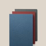 The Layflat Notebook Bundle (Series 2 - Set of 3) features a blue notebook with an embossed geometric design, a red one, and a dark gray one, each with rounded corners and stacked slightly offset on a light beige background.