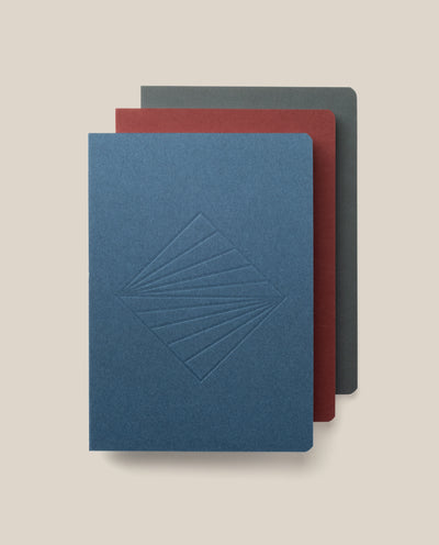 The Layflat Notebook Bundle (Series 2 - Set of 3) features a blue notebook with an embossed geometric design, a red one, and a dark gray one, each with rounded corners and stacked slightly offset on a light beige background.