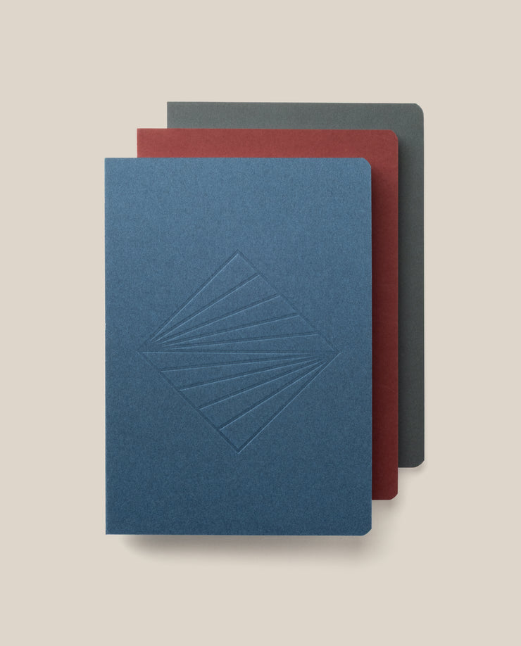 The Layflat Notebook Bundle (Series 2 - Set of 3) features a blue notebook with an embossed geometric design, a red one, and a dark gray one, each with rounded corners and stacked slightly offset on a light beige background.
