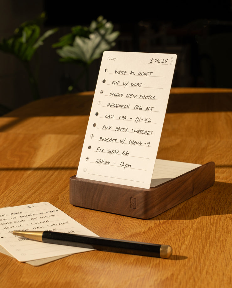 The Analog Daily Focus Kit (Walnut) rests on a wooden table beside a pencil and task cards, with sunlight casting warm shadows that accentuate this simple yet effective productivity system.