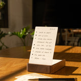 An Analog Annual Kit (Walnut) sits on a sunlit wooden table, displaying a to-do list on its card holder. Nearby are a pen, an extra notepad page, and green plants under soft indoor lighting in the background.