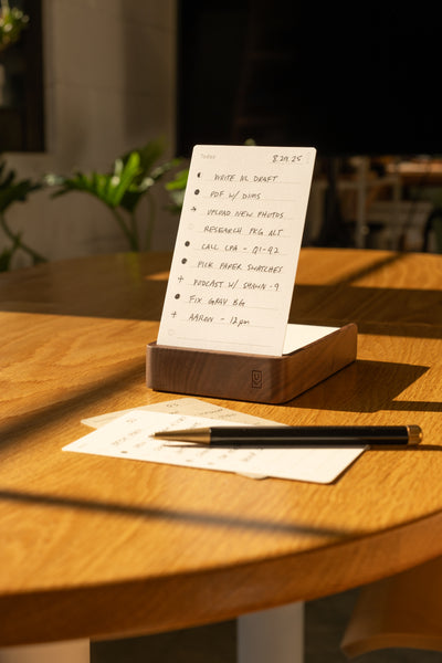 An Analog Annual Kit (Walnut) sits on a sunlit wooden table, displaying a to-do list on its card holder. Nearby are a pen, an extra notepad page, and green plants under soft indoor lighting in the background.