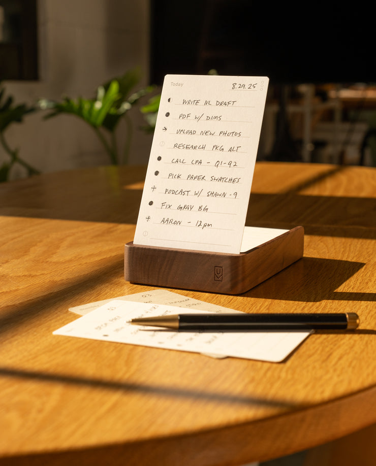 An Analog Annual Kit (Walnut) sits on a sunlit wooden table, displaying a to-do list on its card holder. Nearby are a pen, an extra notepad page, and green plants under soft indoor lighting in the background.