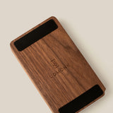 A rectangular walnut Analog Annual Kit with two black strips at the top and bottom, engraved with the Ugmonk logo and name in the center, photographed on a light beige background.