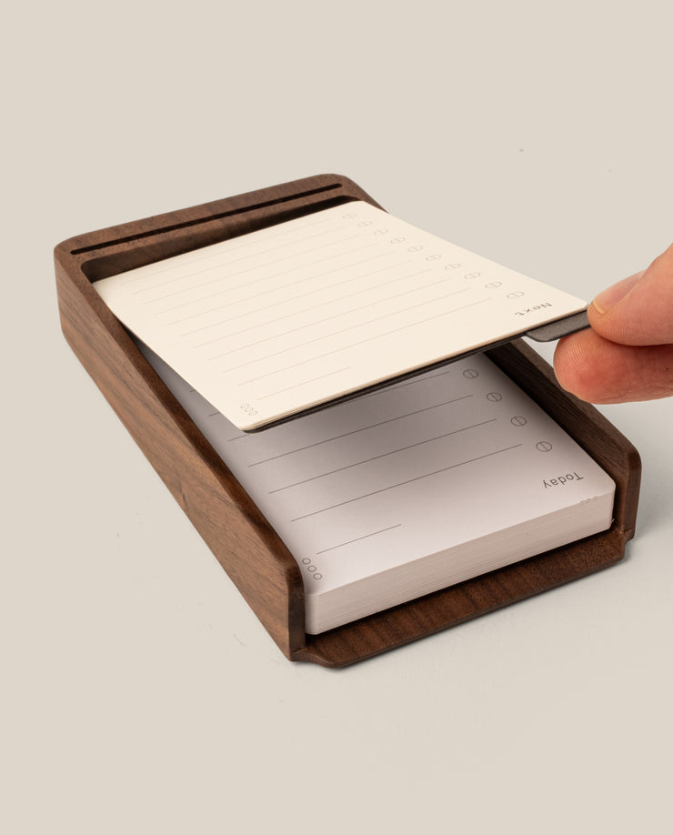 A hand lifts a lined notepad sheet from the Analog Annual Kit (Walnut), a wooden desk organizer designed as a Daily and Weekly Card Holder with checkboxes and lines for notes or tasks.