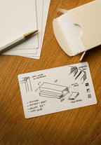 A dotted note card with hand-drawn woodworking plans, measurements, and labels sits on a wooden table, next to a pen and an open box of similar note cards.