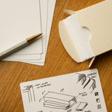 A dotted note card with hand-drawn woodworking plans, measurements, and labels sits on a wooden table, next to a pen and an open box of similar note cards.