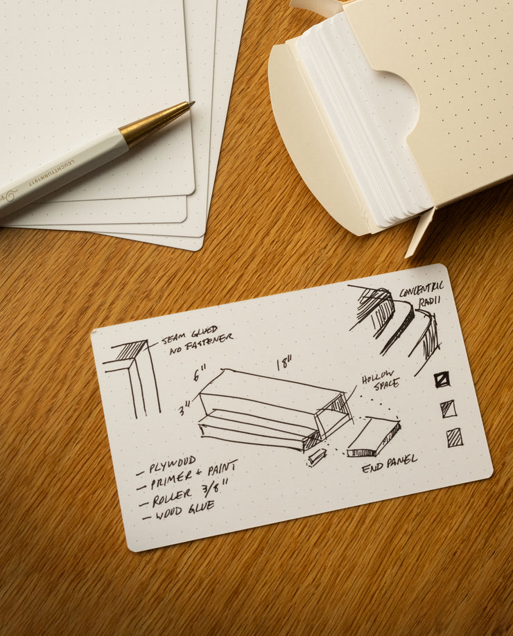 A dotted note card with hand-drawn woodworking plans, measurements, and labels sits on a wooden table, next to a pen and an open box of similar note cards.