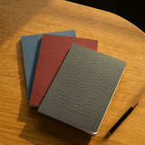 The Layflat Notebook Bundle (Series 2 - Set of 3) sits stacked on a wooden surface beside a black and gold pen, with sunlight casting soft shadows across the table.