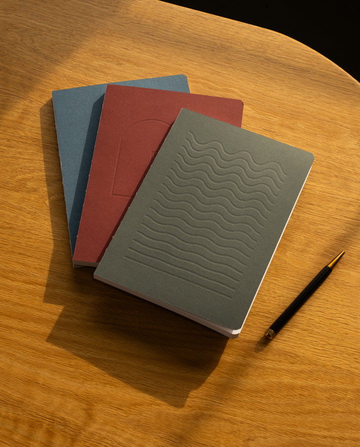 The Layflat Notebook Bundle (Series 2 - Set of 3) sits stacked on a wooden surface beside a black and gold pen, with sunlight casting soft shadows across the table.
