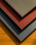 A stack of three Layflat Notebooks (Charcoal) with textured covers and Smyth sewn binding rests on wood, their visible spines highlighted by sunlight casting soft shadows.