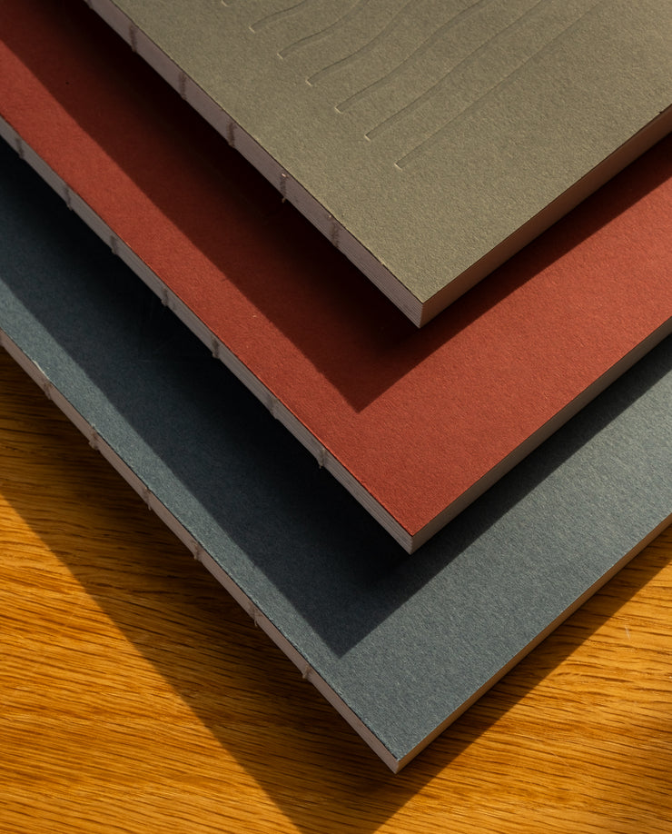 A stack of three Layflat Notebooks (Charcoal) with textured covers and Smyth sewn binding rests on wood, their visible spines highlighted by sunlight casting soft shadows.