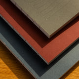 The Layflat Notebook Bundle (Series 2 - Set of 3) features gray, red, and teal dot grid notebooks with exposed Smyth sewn binding, resting on a wooden surface where sunlight highlights their textured paper and covers.