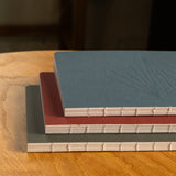 A Layflat Notebook (Maroon) with visible beige spine stitching sits on a wooden table. Its maroon cover features a subtle embossed design, making it a stylish dot grid notebook—perfect for notes or journaling.