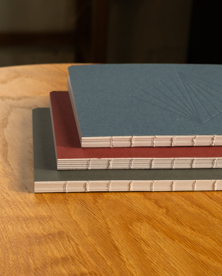 A Layflat Notebook (Maroon) with visible beige spine stitching sits on a wooden table. Its maroon cover features a subtle embossed design, making it a stylish dot grid notebook—perfect for notes or journaling.
