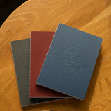 The Layflat Notebook Bundle (Series 2 - Set of 3) features textured gray, maroon, and blue notebooks arranged in a fan shape on a wooden table, with a black and gold pen placed below them.
