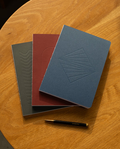 The Layflat Notebook Bundle (Series 2 - Set of 3) features textured gray, maroon, and blue notebooks arranged in a fan shape on a wooden table, with a black and gold pen placed below them.