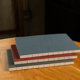 The Layflat Notebook Bundle (Series 2 - Set of 3) features three layflat notebooks with visible spine stitching and plain blue, red, and gray covers, stacked on a round wooden table. The top notebook’s cover includes a geometric embossed design.