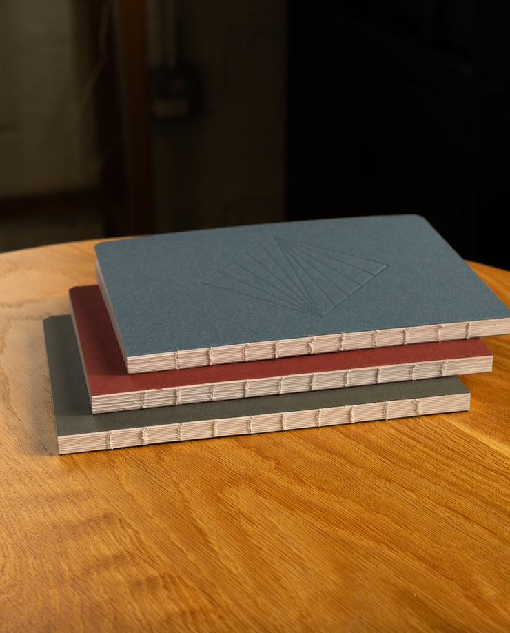 The Layflat Notebook Bundle (Series 2 - Set of 3) features three layflat notebooks with visible spine stitching and plain blue, red, and gray covers, stacked on a round wooden table. The top notebook’s cover includes a geometric embossed design.