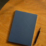 A closed Layflat Notebook (Navy) with a geometric embossed design rests on a wooden surface beside a black and gold pen.