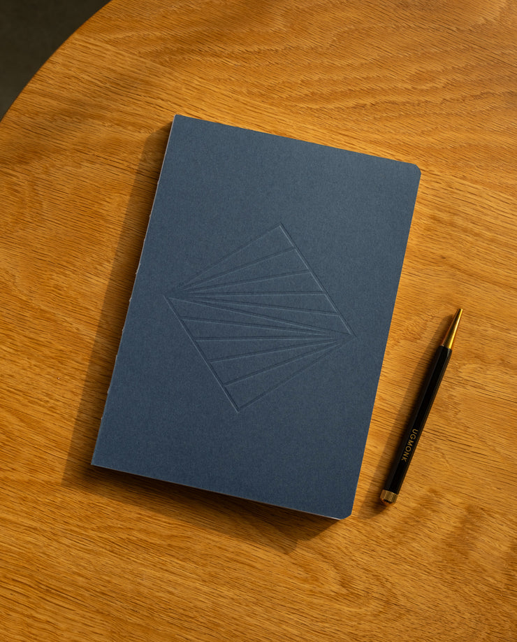 A closed Layflat Notebook (Navy) with a geometric embossed design rests on a wooden surface beside a black and gold pen.
