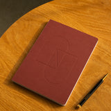 A closed Layflat Notebook (Maroon) rests on a round wooden table next to a black and gold pen.