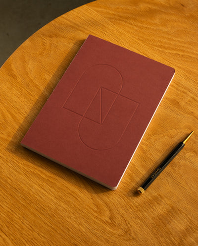 A closed Layflat Notebook (Maroon) rests on a round wooden table next to a black and gold pen.