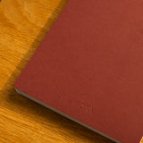 A Layflat Notebook (Maroon) with “UGMONK” embossed on the cover rests on a wooden surface.