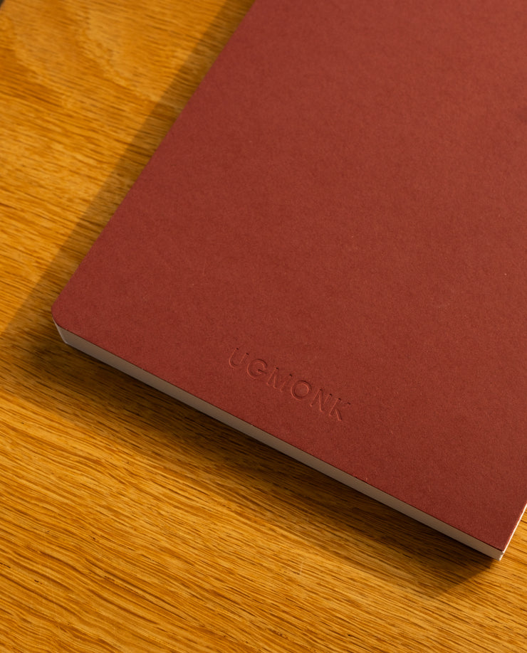 A Layflat Notebook (Maroon) with “UGMONK” embossed on the cover rests on a wooden surface.