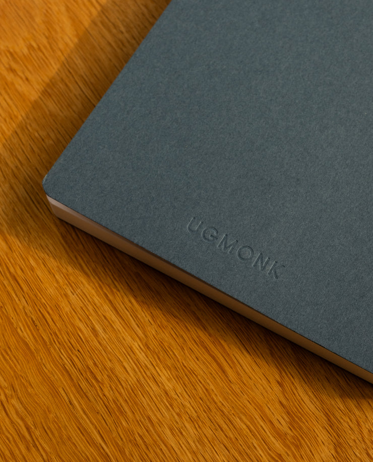 A close-up of a navy Layflat Notebook with dot grid pages and Smyth sewn binding, featuring the UGMONK debossed logo on the cover, resting on a wooden surface.
