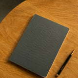 A closed Layflat Notebook (Charcoal) with wavy embossed lines rests on a round wooden table beside a diagonally placed black and gold pen.