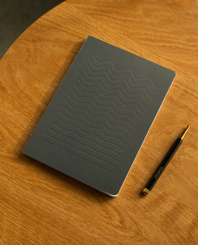 A closed Layflat Notebook (Charcoal) with wavy embossed lines rests on a round wooden table beside a diagonally placed black and gold pen.