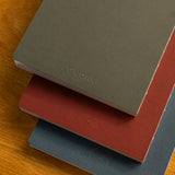 The Layflat Notebook Bundle (Series 2 - Set of 3) features minimalist olive green, rusty red, and navy blue notebooks, each embossed with UGMONK, displayed stacked on a wooden surface.