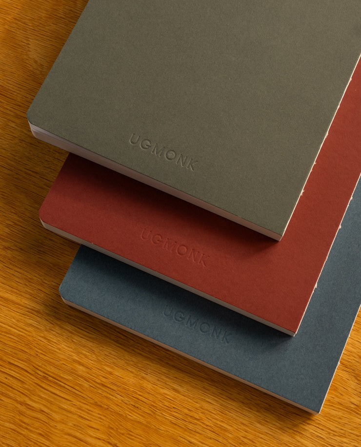 The Layflat Notebook Bundle (Series 2 - Set of 3) features minimalist olive green, rusty red, and navy blue notebooks, each embossed with UGMONK, displayed stacked on a wooden surface.