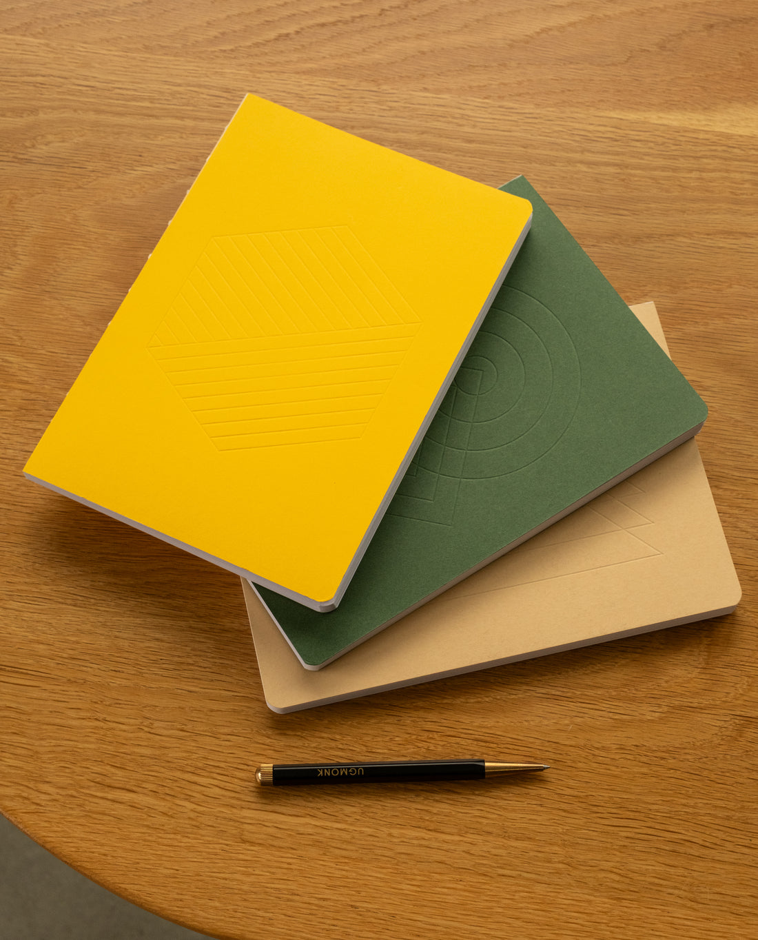 Three notebooks with yellow, green, and beige covers are stacked on a wooden surface. A black pen lies below the notebooks. The covers have subtle geometric embossed designs.
