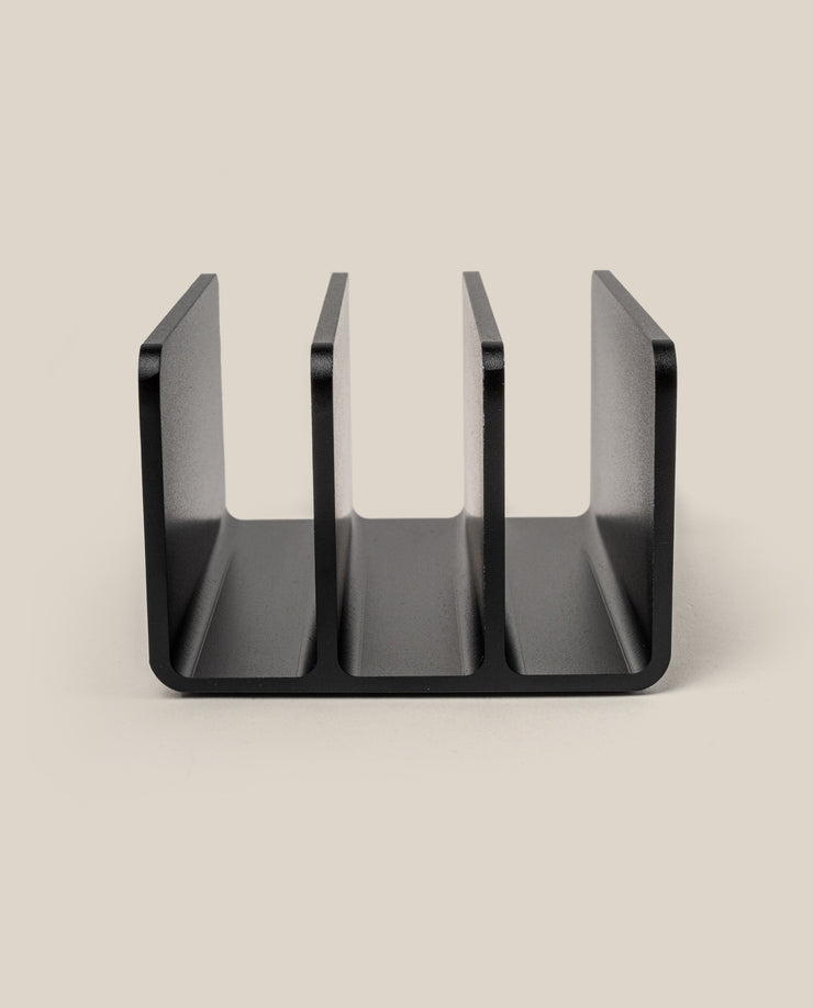 A black, three-slot desktop organizer from The Complete Analog Collection (Black Aluminum) stands upright on a beige surface, photographed from the front to display its U-shaped, open compartments.