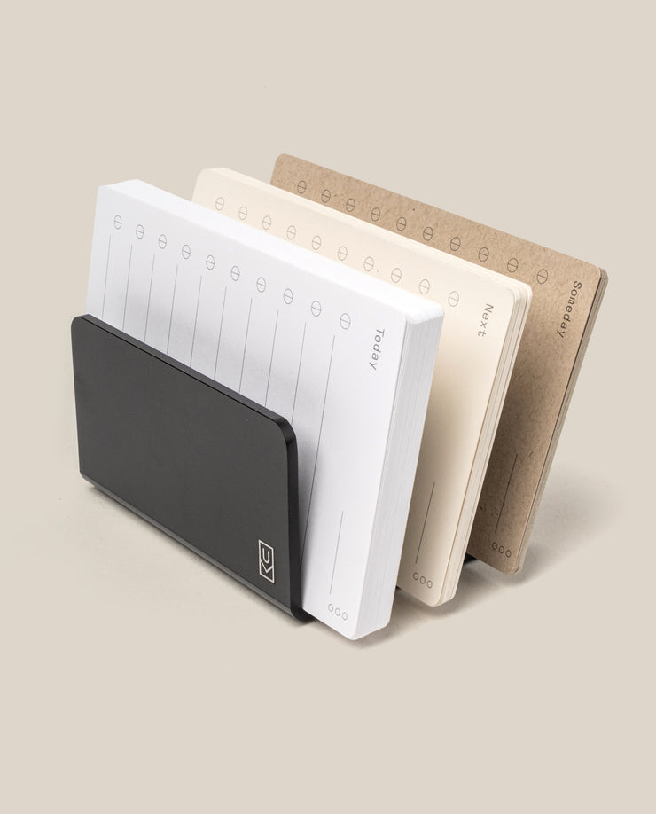 The Complete Analog Collection (Black Aluminum): a black desktop organizer with three notepads—white, cream, and brown—for weekly planning in a horizontal layout, shown against a neutral beige background.