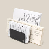 Three open notebooks with handwritten notes and sketches are arranged upright with the Analog Card Organizer (Black Aluminum) leaning against them, all set against a plain beige background.