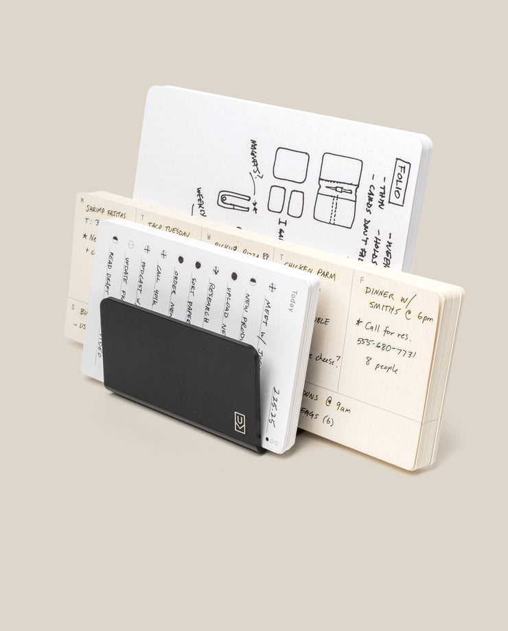 Three open notebooks with handwritten notes and sketches are arranged upright with the Analog Card Organizer (Black Aluminum) leaning against them, all set against a plain beige background.